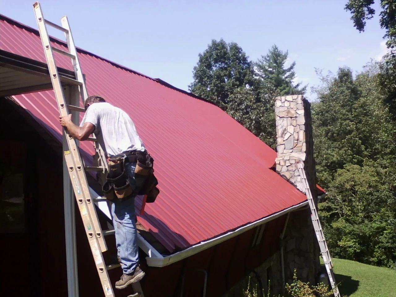 Roofer on a ladder beside a red metal roof and stone chimney — Soffit & Fascia Repair by Roofing Brothers in Heiskell